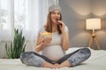Pregnant happy smiling woman sitting on bed eating unhealthy junk food snack young beautiful mom expecting baby enjoying potato Royalty Free Stock Photo