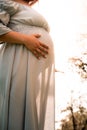 Pregnant Latina woman stands in a park, hands gently cradling her rounded belly