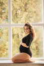 Pregnant moman sits in front of big window. Model dressed in black bodysuit. She put her hand on the belly. Happy future Royalty Free Stock Photo