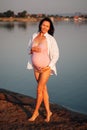 a pregnant woman on the beach, Smiling, dark-haired pregnant woman in a pink swimsuit and a white shirt stands holding Royalty Free Stock Photo