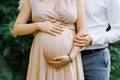 A pregnant woman in a beige long dress holds her hands on her stomach as her husband and baby daddy-to-be hugs his wife. Close-up