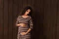 Pregnant woman in brown stripe dress holds hands on belly on a dark brown background. Pregnancy, maternity, preparation and expect