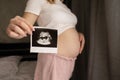 Pregnant woman demonstrating her ultrasound photo while standing at bedroom. Picture with ultrasound scan result