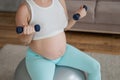 Pregnant woman doing exercises with dumbbells while sitting on a fitness ball at home. Close-up of a pregnant belly. Royalty Free Stock Photo