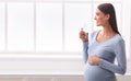Pregnant Woman Drinking Glass Of Water Standing Against Window Indoor