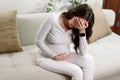 Pregnant woman experiences pain, holds belly with hands sitting on couch, discomfort, headache