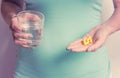 A pregnant woman first trimester in a blue T-shirt holds several medicine capsules, and in the second a glass of water. IVF.