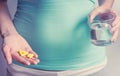 A pregnant woman first trimester in a blue T-shirt holds several medicine capsules, and in the second a glass of water. IVF.