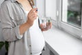 Pregnant woman holding pill and glass of water near window indoors, closeup Royalty Free Stock Photo