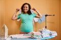 Pregnant woman holds newborn clothes and steam iron, smiles at camera standing at ironing board with ironed baby clothes Royalty Free Stock Photo