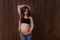 Pregnant woman in jeans and black top holds hands on belly on a dark brown background. Pregnancy, maternity, preparation and