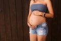 Pregnant woman in jeans shorts and blue top holds hands on belly on a dark brown background. Pregnancy, maternity. Beautiful tende Royalty Free Stock Photo