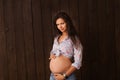 Pregnant woman in jeans shorts and blue top holds hands on belly on a dark brown background. Pregnancy, maternity, preparation and Royalty Free Stock Photo