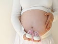 Pregnant woman with long hair in jeans on a white background holds in the hands near the belly baby booties in the room at home. Royalty Free Stock Photo