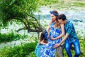 Pregnant woman mother to be in wreath and blue dress sitting with her daughter and husband near scenic river in sunset time in Royalty Free Stock Photo