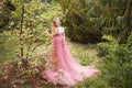 Pregnant woman in pink long dress standing in blooming magnolia in forest