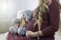 Pregnant woman in red dress sitting comfortably on a sofa. Her hand is on her belly and with the other she is holding a grey teddy Royalty Free Stock Photo