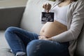 Pregnant woman showing baby sonography photo while sitting on couch at home