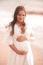 Pregnant woman showing heart shape with fingers wearing white dress over sea background. Motherhood. Maternity. Royalty Free Stock Photo