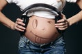 Pregnant woman showing her tummy with smile and holding earphones.