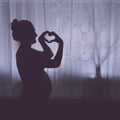 Pregnant woman shows her heart with a hand gesture in the light of a night window, black and white silhouette Royalty Free Stock Photo