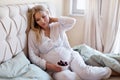 Pregnant woman sitting on bed and eating cherry berries. Thoughtful blonde model in pajamas under blanket touching neck