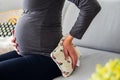 Pregnant woman sitting comfortably on the sofa and holding a therapeutic heat pillow on her lower back