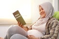 Pregnant woman sitting on couch her belly and reading baby name book. Expecting mother relaxing, preparing name for her baby Royalty Free Stock Photo