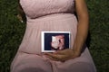 Pregnant woman sitting in the park on the grass holding 3D  image ultrasound of baby in her womb Royalty Free Stock Photo