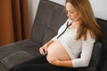 Pregnant woman sitting on sofa and listening to her baby heart-rate with stethoscope. Horizontal shape