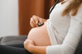 Pregnant woman sitting on sofa and listening to her baby heart-rate with stethoscope. Horizontal shape