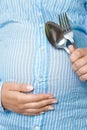 A pregnant woman with a spoon and a fork in her hands is standing on a white background. Diet for a baby in the stomach who will