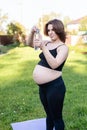 Pregnant woman in sportswear drinking a bottle of water after doing yoga outdoors, taking break in yoga practice