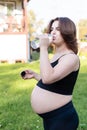 Pregnant woman in sportswear drinking a bottle of water after doing yoga outdoors, taking break in yoga practice