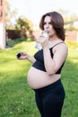 Pregnant woman in sportswear drinking a bottle of water after doing yoga outdoors, taking break in yoga practice