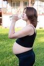 Pregnant woman in sportswear drinking a bottle of water after doing yoga outdoors, taking break in yoga practice