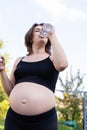 Pregnant woman in sportswear drinking a bottle of water after doing yoga outdoors, taking break in yoga practice