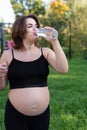 Pregnant woman in sportswear drinking a bottle of water after doing yoga outdoors, taking break in yoga practice