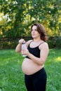 Pregnant woman in sportswear drinking a bottle of water after doing yoga outdoors, taking break in yoga practice