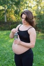 Pregnant woman in sportswear drinking a bottle of water after doing yoga outdoors, taking break in yoga practice