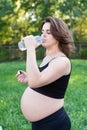 Pregnant woman in sportswear drinking a bottle of water after doing yoga outdoors, taking break in yoga practice