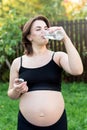 Pregnant woman in sportswear drinking a bottle of water after doing yoga outdoors, taking break in yoga practice