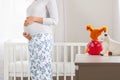 A pregnant woman stands in the children& x27;s white room and hugs her tummy. In the background, a large window and a cradle Royalty Free Stock Photo