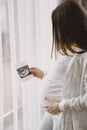 Pregnant woman stands by the window and looks at photo