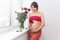 Pregnant woman stands by the window next to a bouquet of roses in a vase