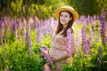 A pregnant woman in a summer dress with a flower crown with a large luxurious blue lupin sits in a field of blooming lupin