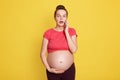 Pregnant woman talking by mobile phone while standing isolated over yellow background, wearing red t shirt, touching her bare Royalty Free Stock Photo