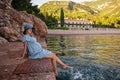 Pregnant woman traveler in denim dress, blue straw hat sits by the sea, dangles legs in water