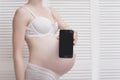 Pregnant woman in underwear holding and showing smartphone with empty display on white wooden screen background. Close-up Royalty Free Stock Photo