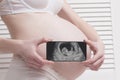 Pregnant woman in underwear holding and showing smartphone with fetal ultrasound photo on white wooden screen background. Close-up